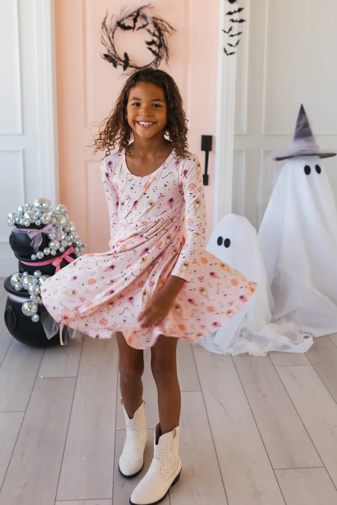 Young girl in a Halloween twirl dress standing in a room decorated for Halloween with a ghost and witch hat.