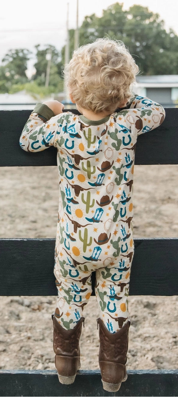 Child wearing patterned howdy romper standing on a fence with a rural background.