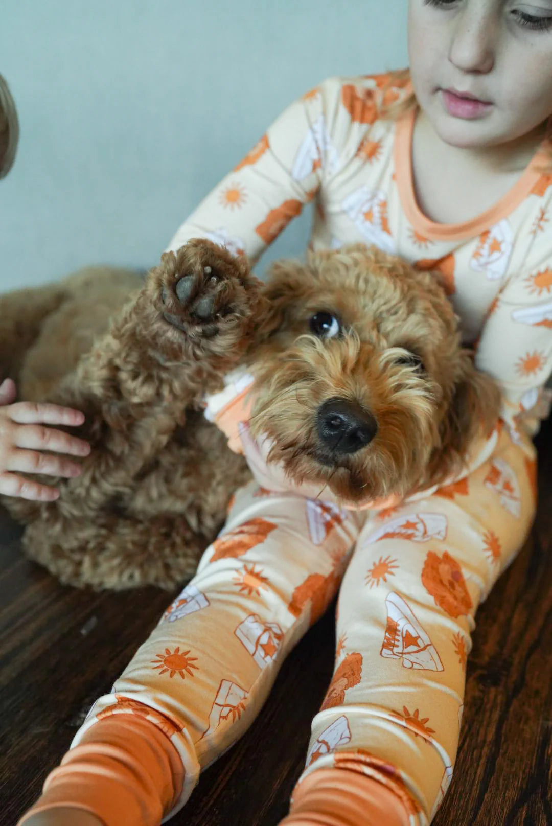 Toddler cuddling goldendoodle puppy while wearing stay golden bamboo romper pajamas. Pajamas are Orange with images of goldendoodles, sneakers and a sun. 