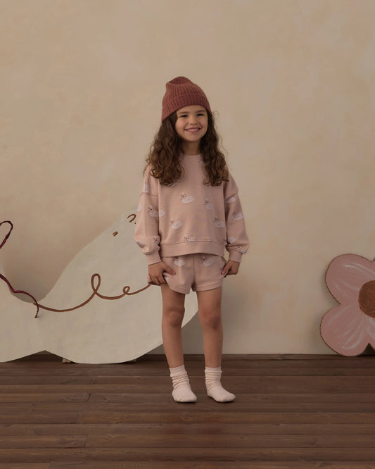 Girl in a rust beanie wearing the Rylee + Cru swans boxy pullover with matching track shorts in front of painted flower cutouts