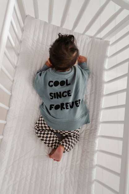 Baby boy laying down in crib wearing our checkered joggers and boxy cool kid sweatshirt. The back of the sweatshirt says "Cool Since Forever" Our bamboo terry is made with 66% Bamboo 28% Cotton and 5% Spandex. 