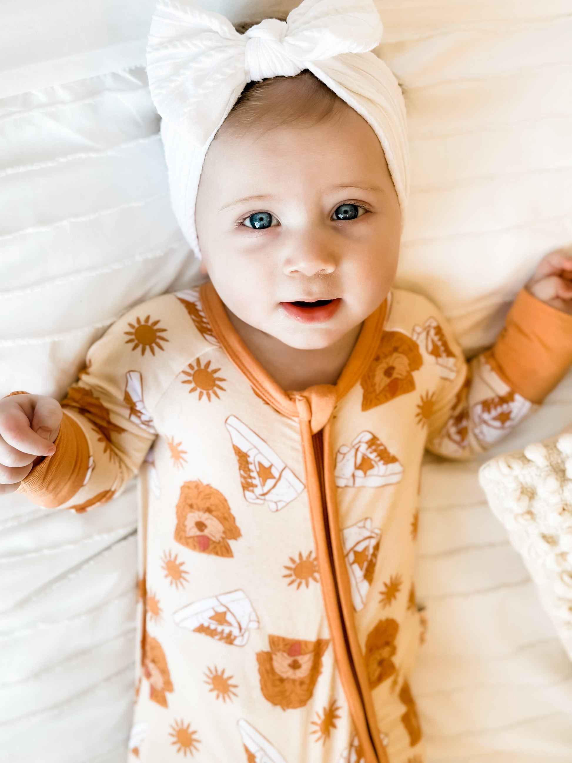 Baby girl laying down while wearing stay golden bamboo romper pajamas. Pajamas are Orange with images of goldendoodles, sneakers and a sun. 